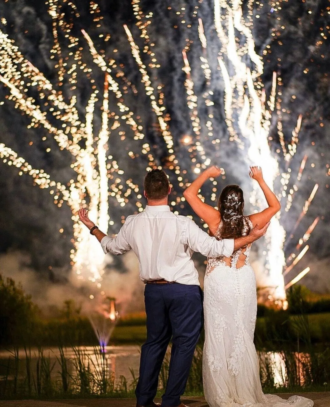 A couple with arms raised under fireworks over the lake